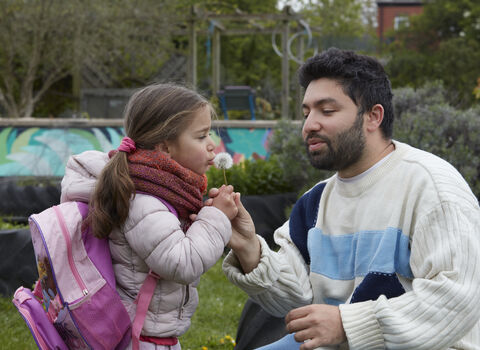A man helps a young girl hold a dandelion clock as she blows the seeds